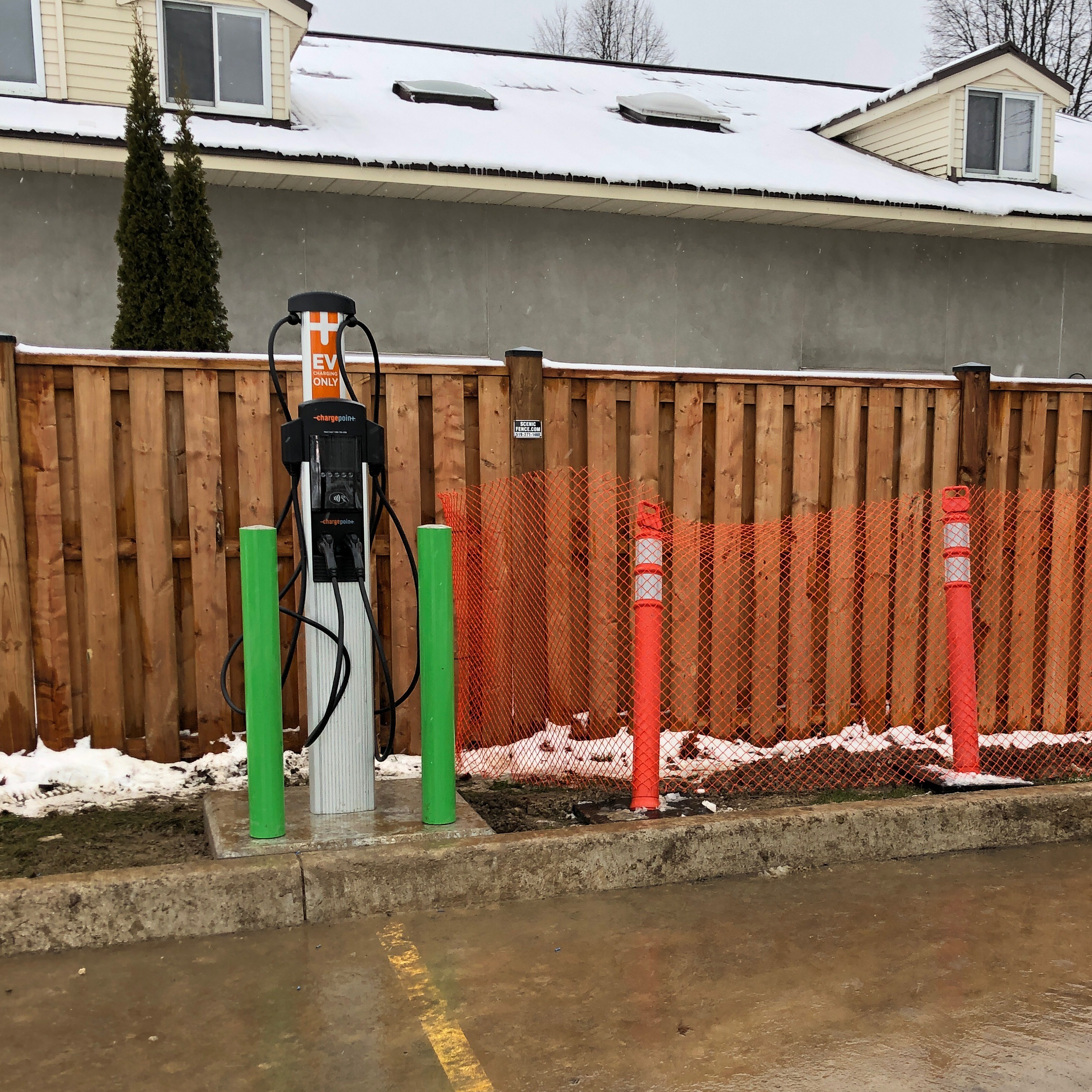 An outdoor ChargePoint electric vehicle charging station with green bollards and orange safety fencing, set against a wooden fence with snow on the ground.