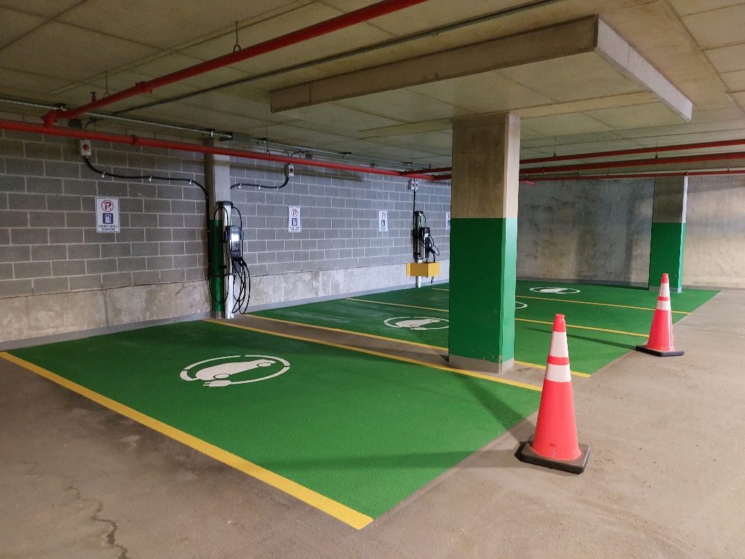 An indoor EV charging area with green pavement painting, ChargePoint chargers mounted on the wall, and orange traffic cones.